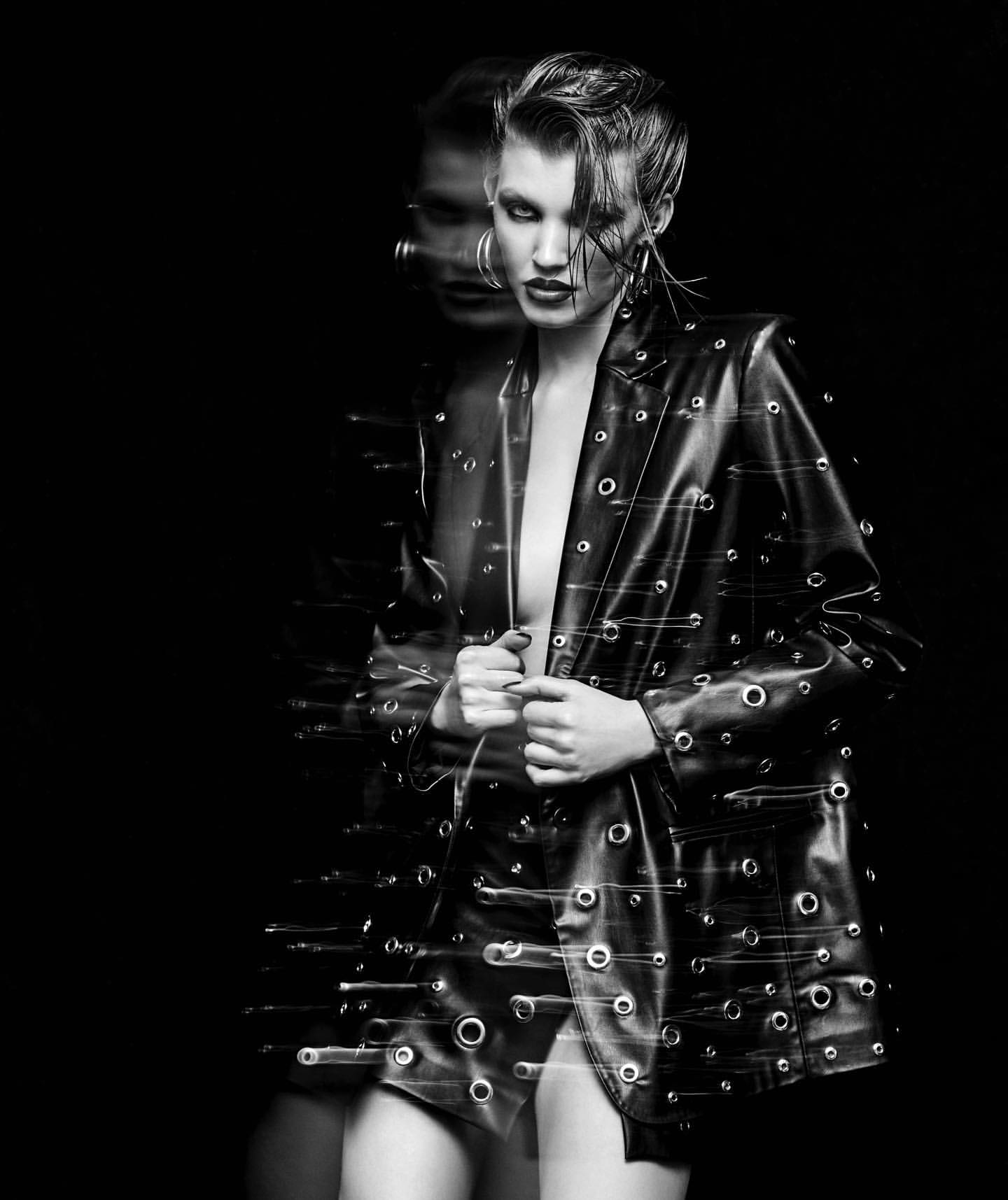Monochrome portrait of a fashionable woman with slicked-back hair in a studded leather jacket against a black background.