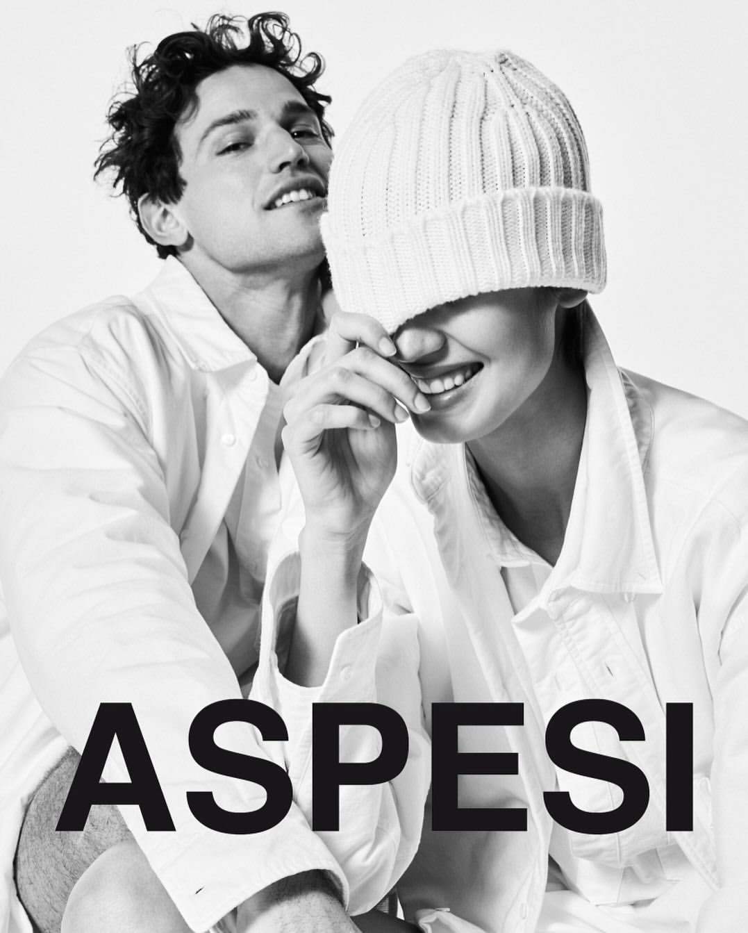 Monochrome fashion shot: a man and woman pose in white jackets, the woman wearing a white knit hat. ASPESI brand logo in the foreground.