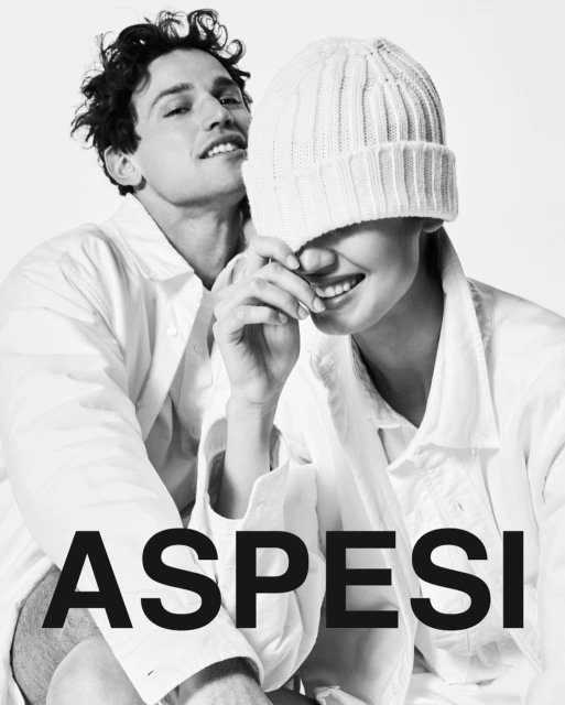 Monochrome fashion shot: a man and woman pose in white jackets, the woman wearing a white knit hat. ASPESI brand logo in the foreground.