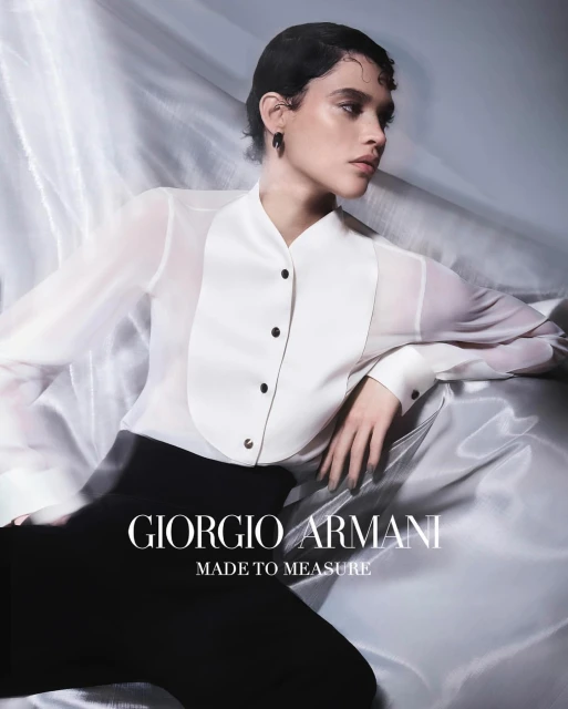 Elegant young woman in sheer white blouse and black pants lying on a silky fabric for a Giorgio Armani fashion advertisement.