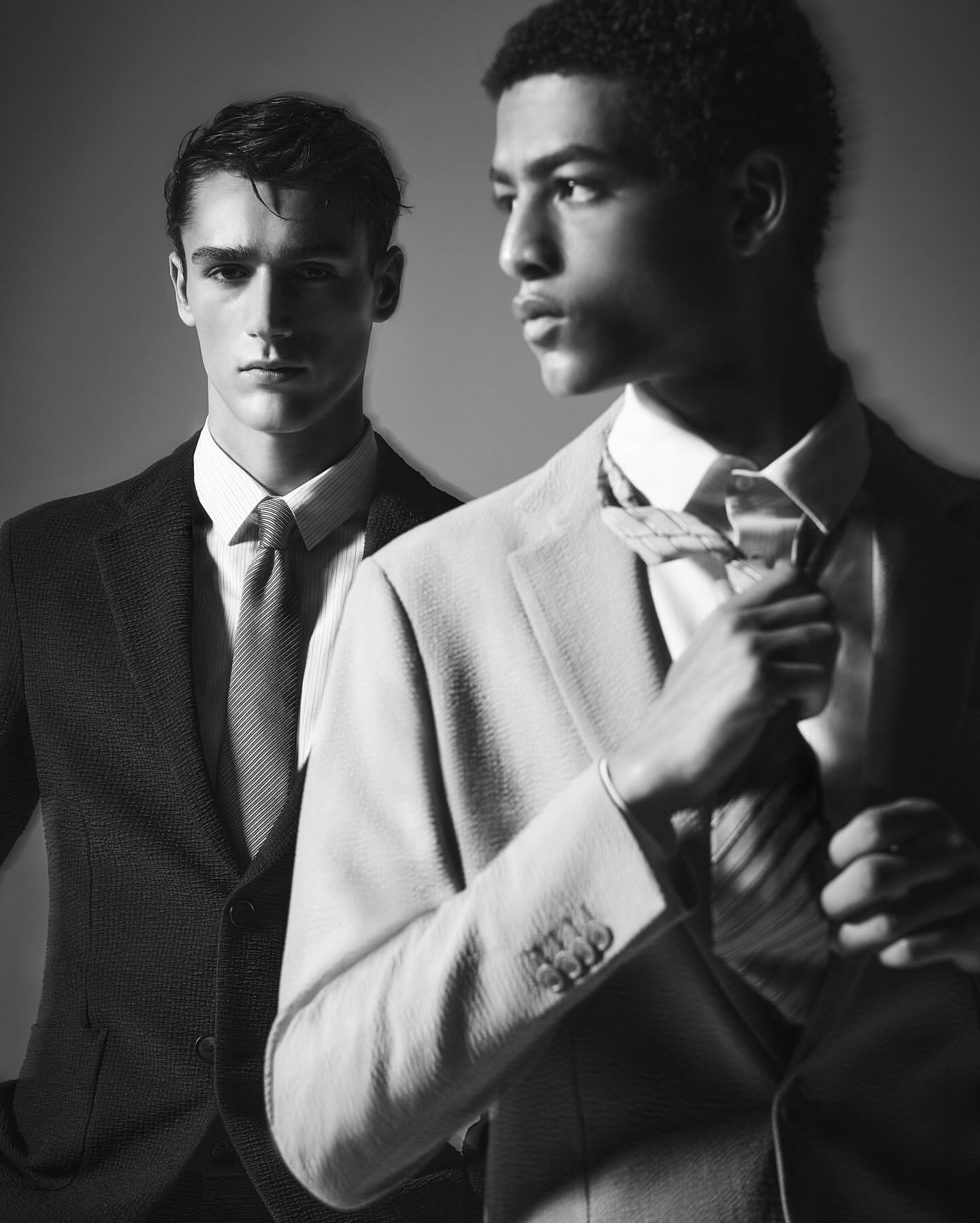 Two men in suits stand indoors, one adjusting his tie, captured in a black and white fashion style portrait.