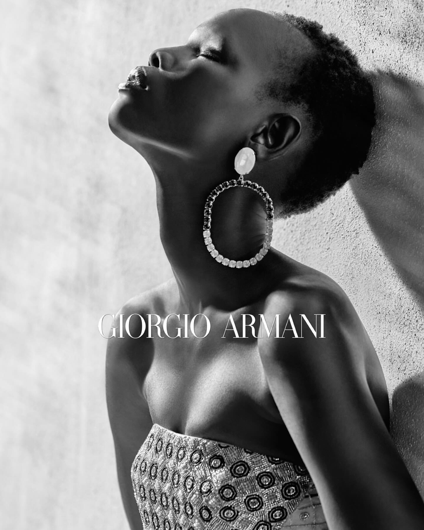 Black and white portrait of a woman wearing large jeweled hoop earrings and a patterned strapless top against a textured wall.