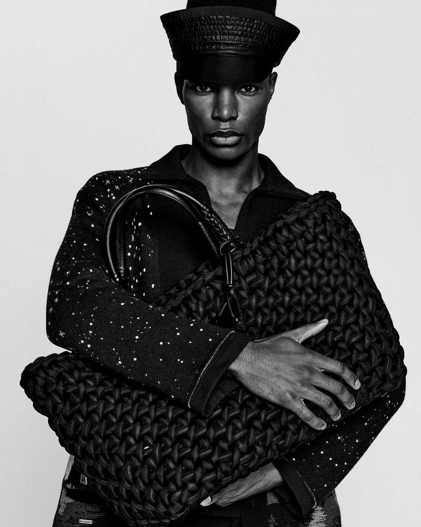 Portrait of an adult male model with dark skin wearing a black outfit and a matching hat, holding a textured black woven handbag indoors.