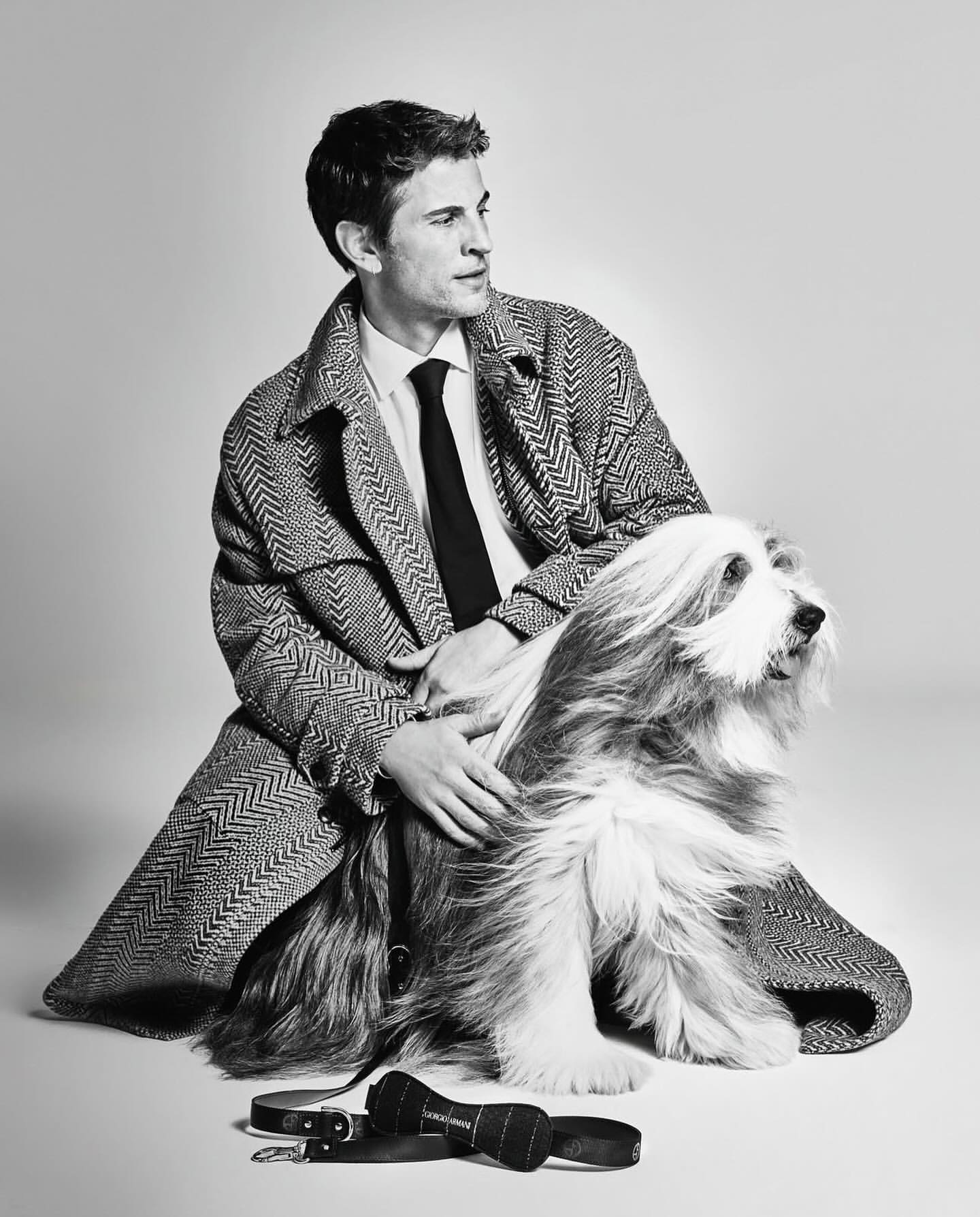 A stylish man in a herringbone coat poses with his bearded collie dog in a studio, showcasing a sophisticated and classic aesthetic.