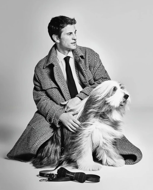 A stylish man in a herringbone coat poses with his bearded collie dog in a studio, showcasing a sophisticated and classic aesthetic.