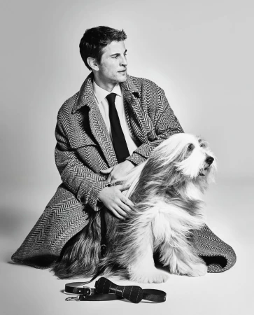 A stylish man in a herringbone coat poses with his bearded collie dog in a studio, showcasing a sophisticated and classic aesthetic.