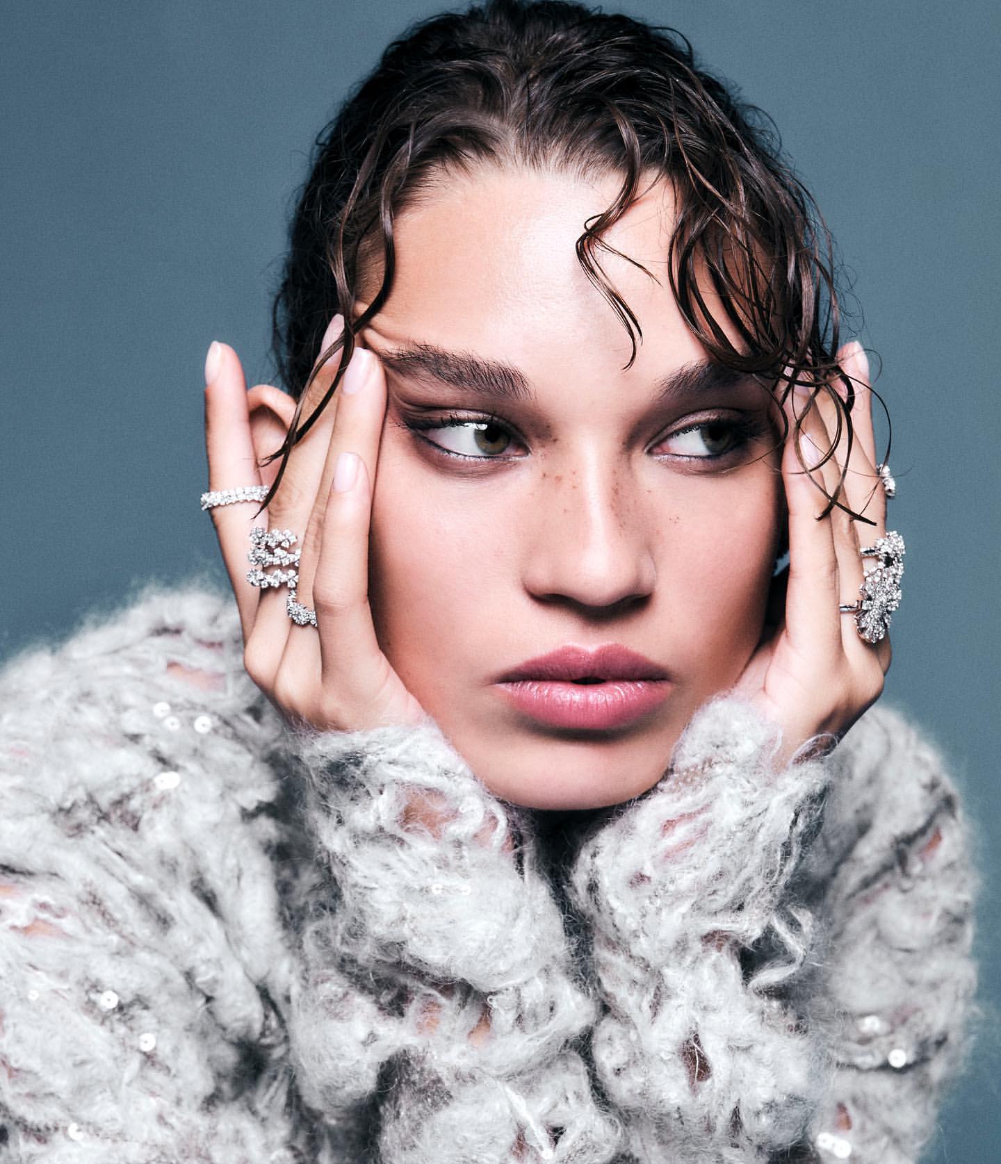 Close-up portrait of a woman with wet hair and jewelry, wearing a fuzzy grey sweater against a neutral blue background.