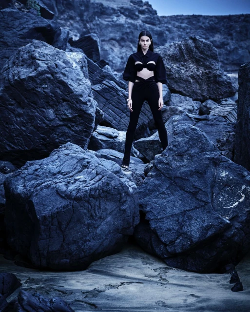 A woman in a dark two-piece outfit standing among dark rocks, looking at the camera, on a blue-tinted day with subdued lighting.