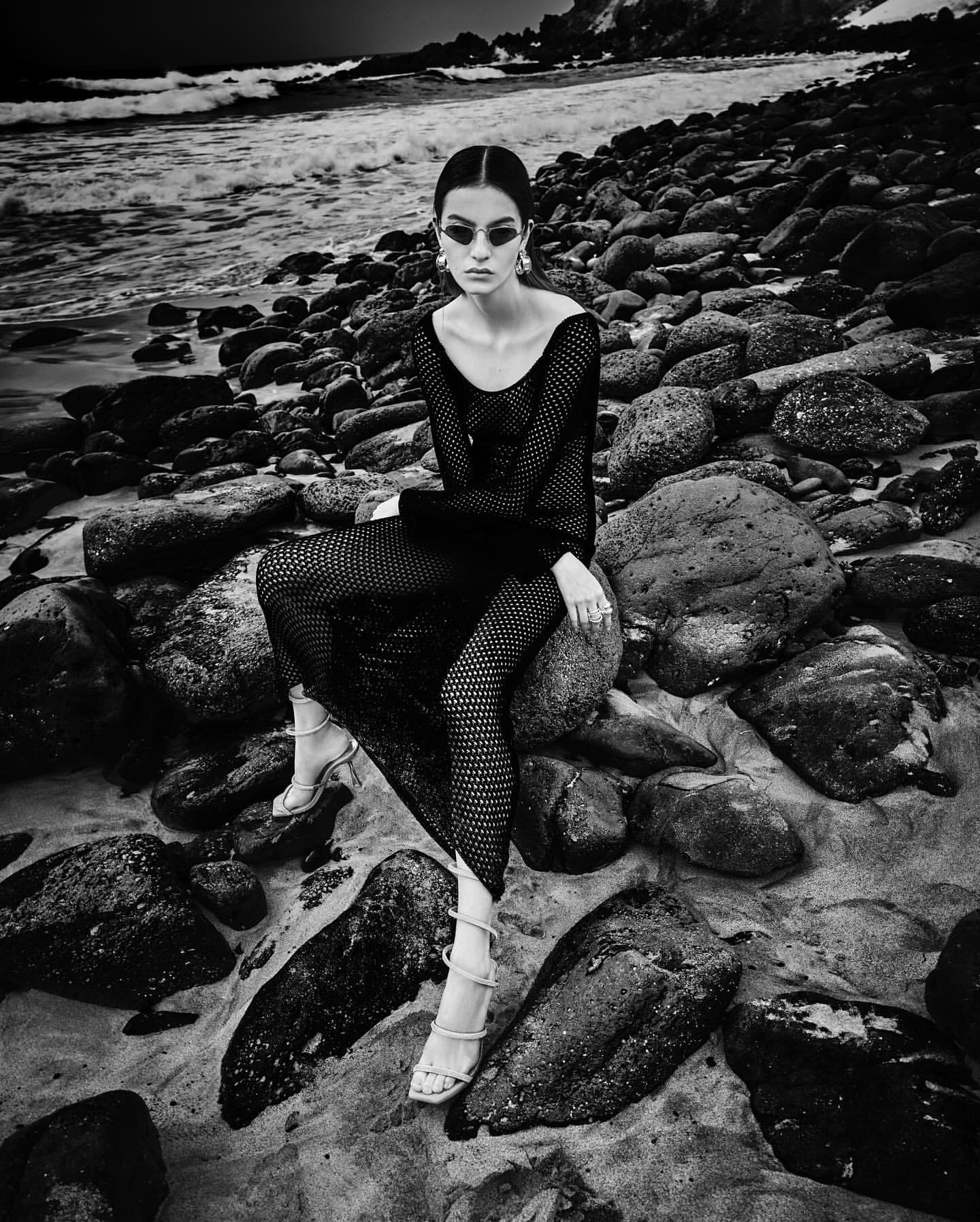 Monochrome fashion portrait of a woman in a net dress, sitting on rocks at the beach with the ocean and sky in the background.