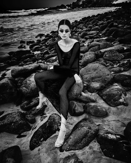 Monochrome fashion portrait of a woman in a net dress, sitting on rocks at the beach with the ocean and sky in the background.