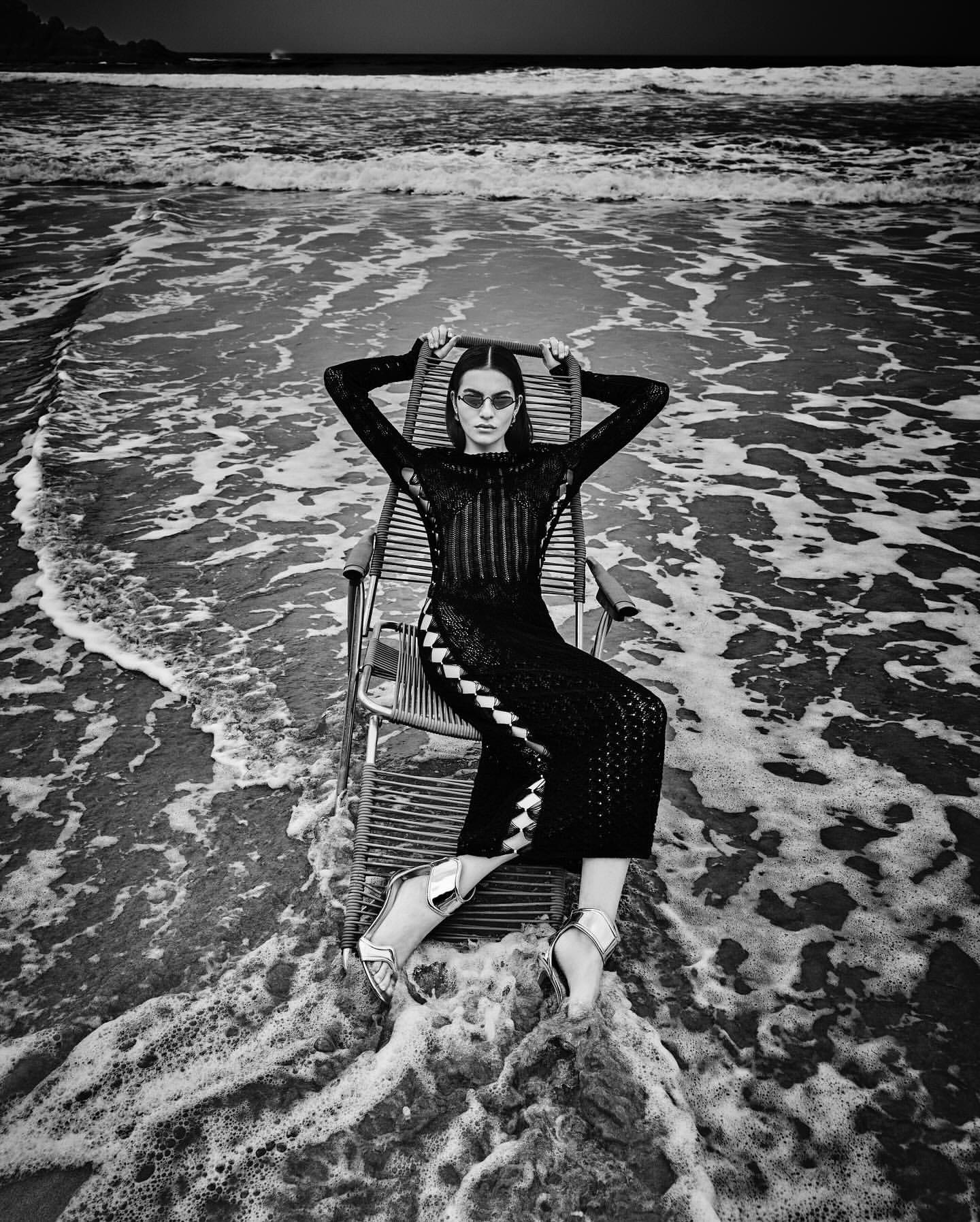 A slender woman in a black dress and sunglasses reclines on a beach chair, the ocean waves washing around her, creating a striking black and white fashion portrait.