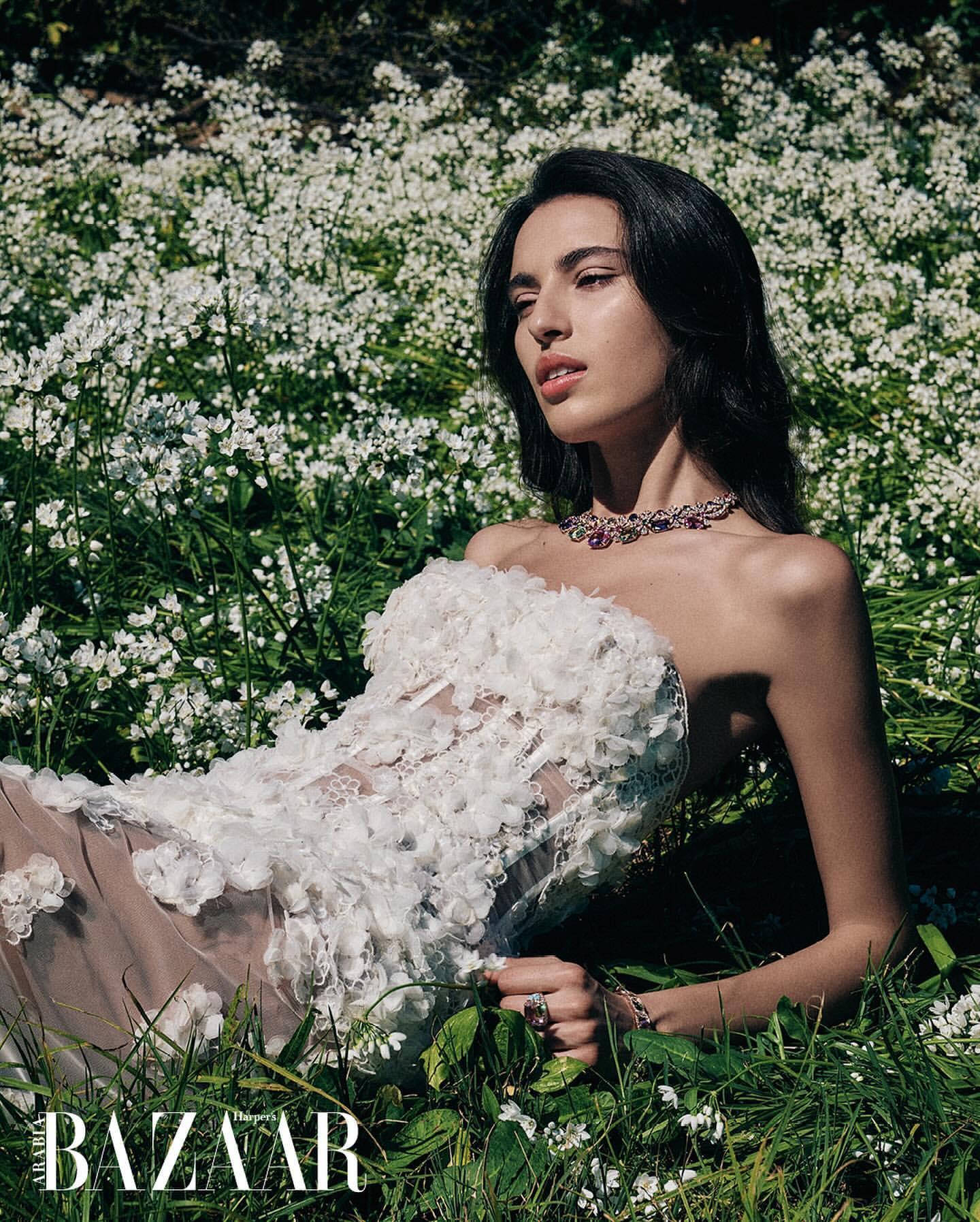 A woman with dark hair and a floral dress lies in a field of white flowers in a fashion editorial shot for Harper's Bazaar magazine.