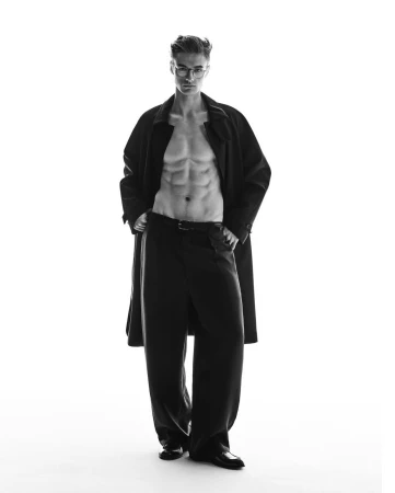 Monochrome portrait of a shirtless man wearing glasses and a long dark coat standing against a plain white background.