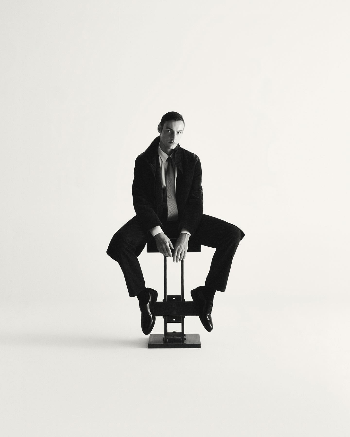 A man in a dark suit and tie seated on a metal stool against a white background by Giulio Rustichelli.