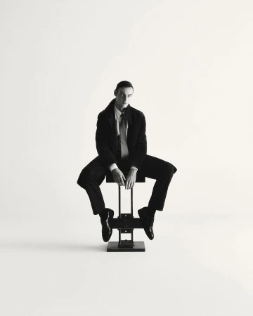 A man in a dark suit and tie seated on a metal stool against a white background by Giulio Rustichelli.