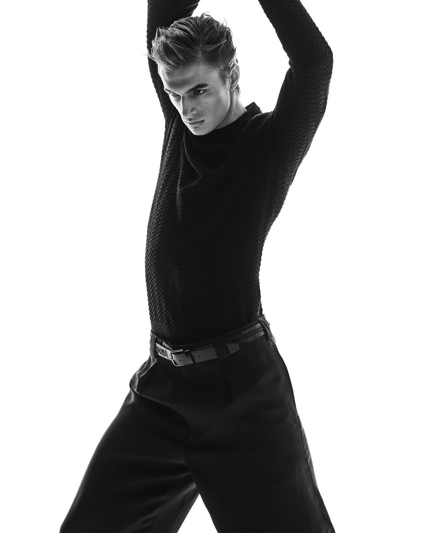 Monochrome portrait of a young adult man with styled hair wearing black textured sweater and trousers against a white background in a studio.