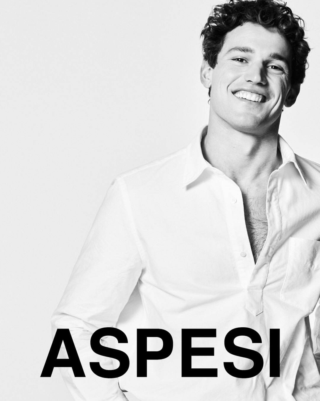 Monochrome portrait of a smiling man in a white button-down shirt against a white seamless backdrop with Aspesi branding