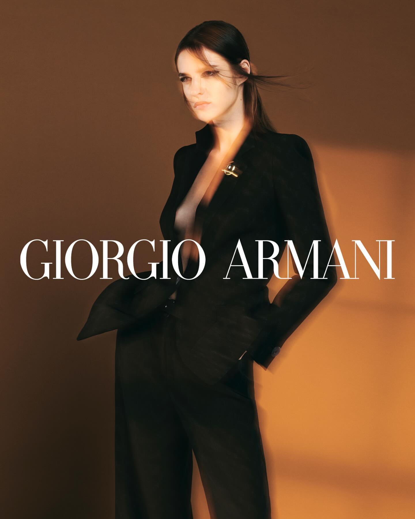 An adult female poses in a black suit with an unbuttoned jacket and wide-leg trousers, set against a warm brown backdrop with the brand logo.