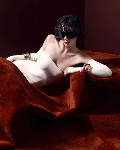 Model in white strapless dress wearing dark sunglasses and thick gold cuffs reclining on a deep rust-colored textured surface.
