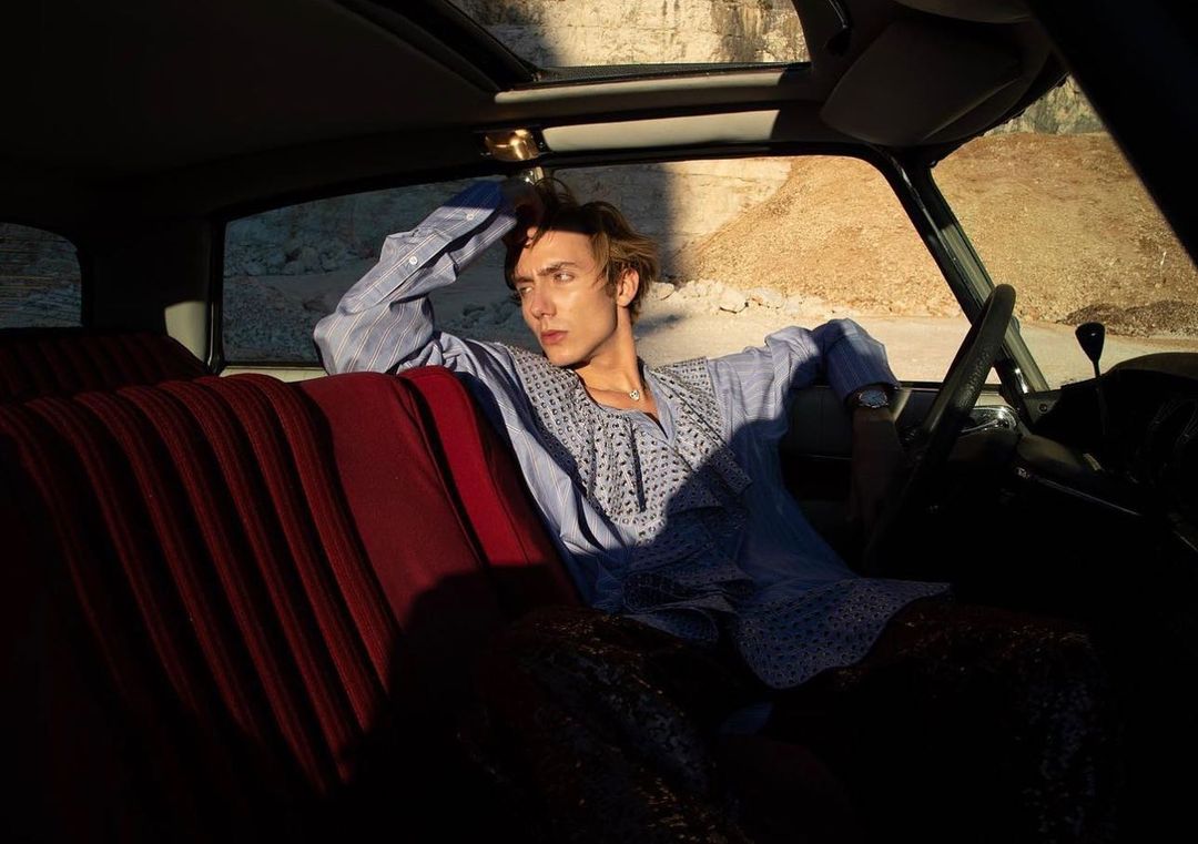 Young adult man in vintage car interior, blue patterned shirt, looking away, with neutral expression, bathed in sunlit golden hour.