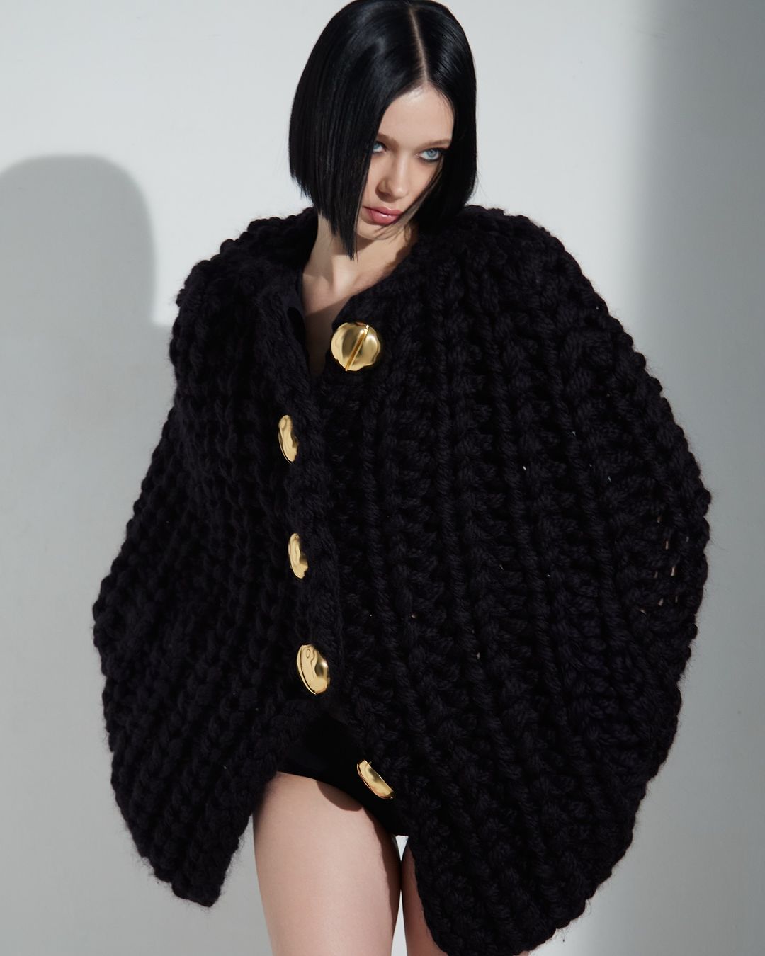 Woman in a chunky black knit cardigan with gold buttons poses against a neutral background in a studio setting. Fashion photography.
