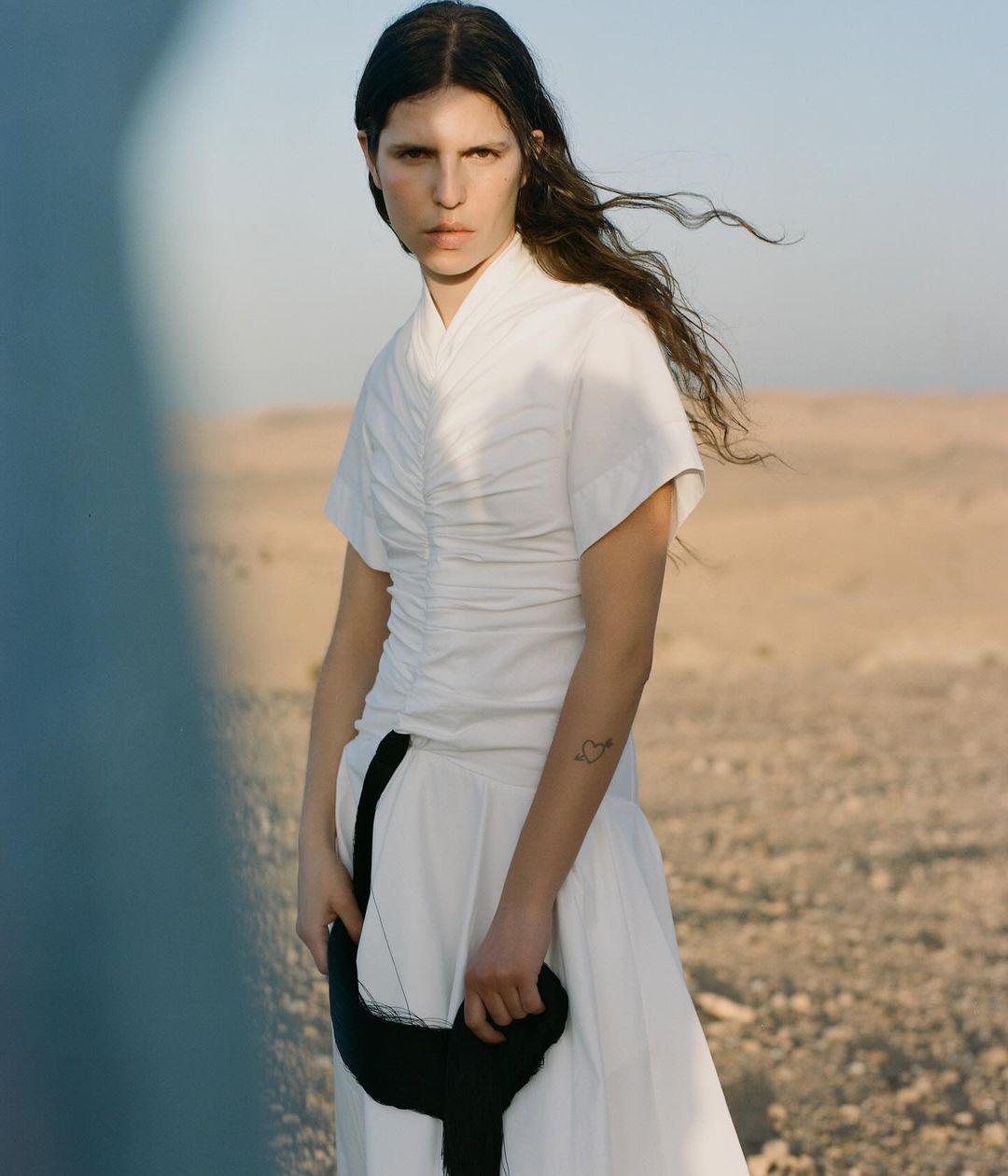 Model in white ruched dress with black strap standing outdoors in a desert landscape photographed by Giuseppe Triscari