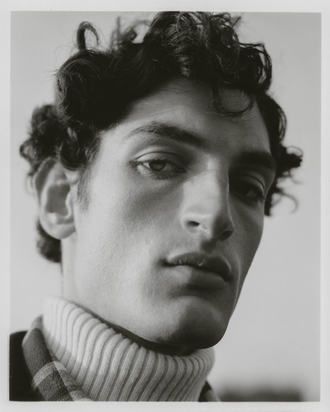 Monochrome portrait of a young adult male with curly dark hair, wearing a turtleneck sweater, in a softly lit, neutral setting.