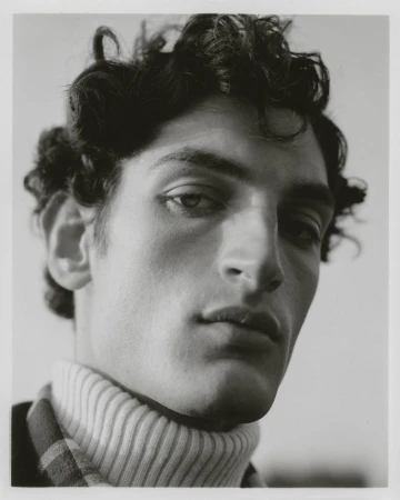 Monochrome portrait of a young adult male with curly dark hair, wearing a turtleneck sweater, in a softly lit, neutral setting.