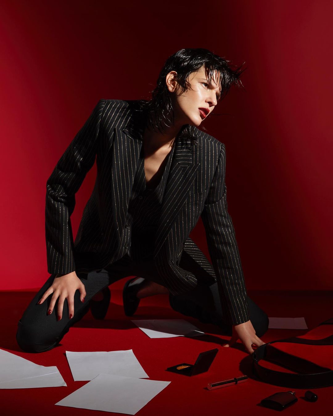 Stylish woman in a pinstripe suit kneels on a red surface surrounded by papers and accessories in a studio setting.
