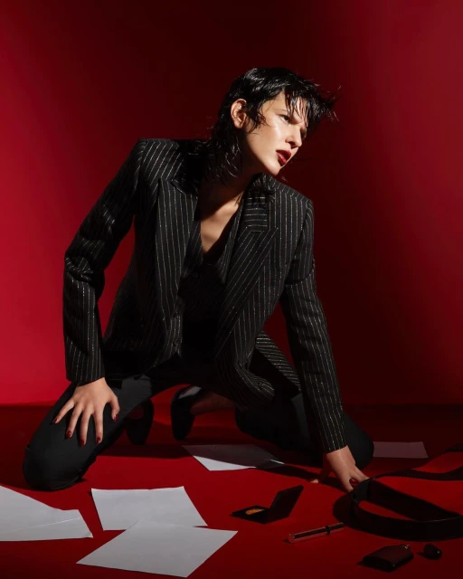 Stylish woman in a pinstripe suit kneels on a red surface surrounded by papers and accessories in a studio setting.
