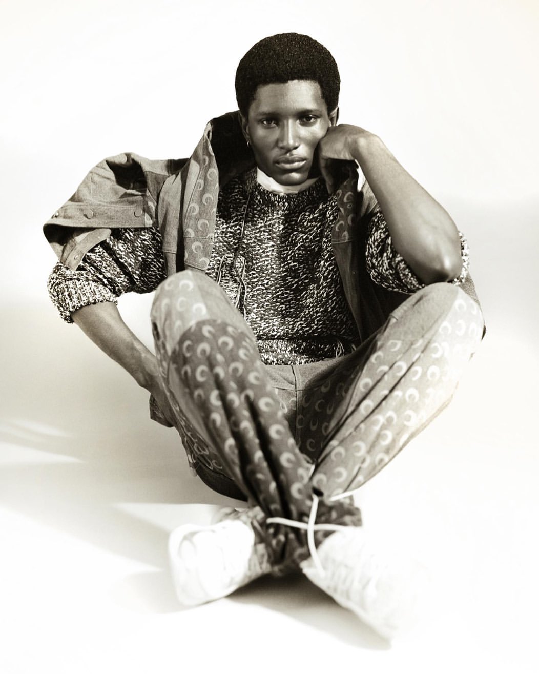 Monochrome studio portrait of a Black man wearing a patterned sweater and trousers, sitting cross-legged against a white backdrop.