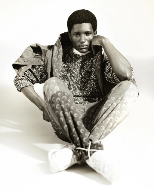 Monochrome studio portrait of a Black man wearing a patterned sweater and trousers, sitting cross-legged against a white backdrop.