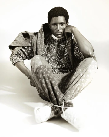 Monochrome studio portrait of a Black man wearing a patterned sweater and trousers, sitting cross-legged against a white backdrop.