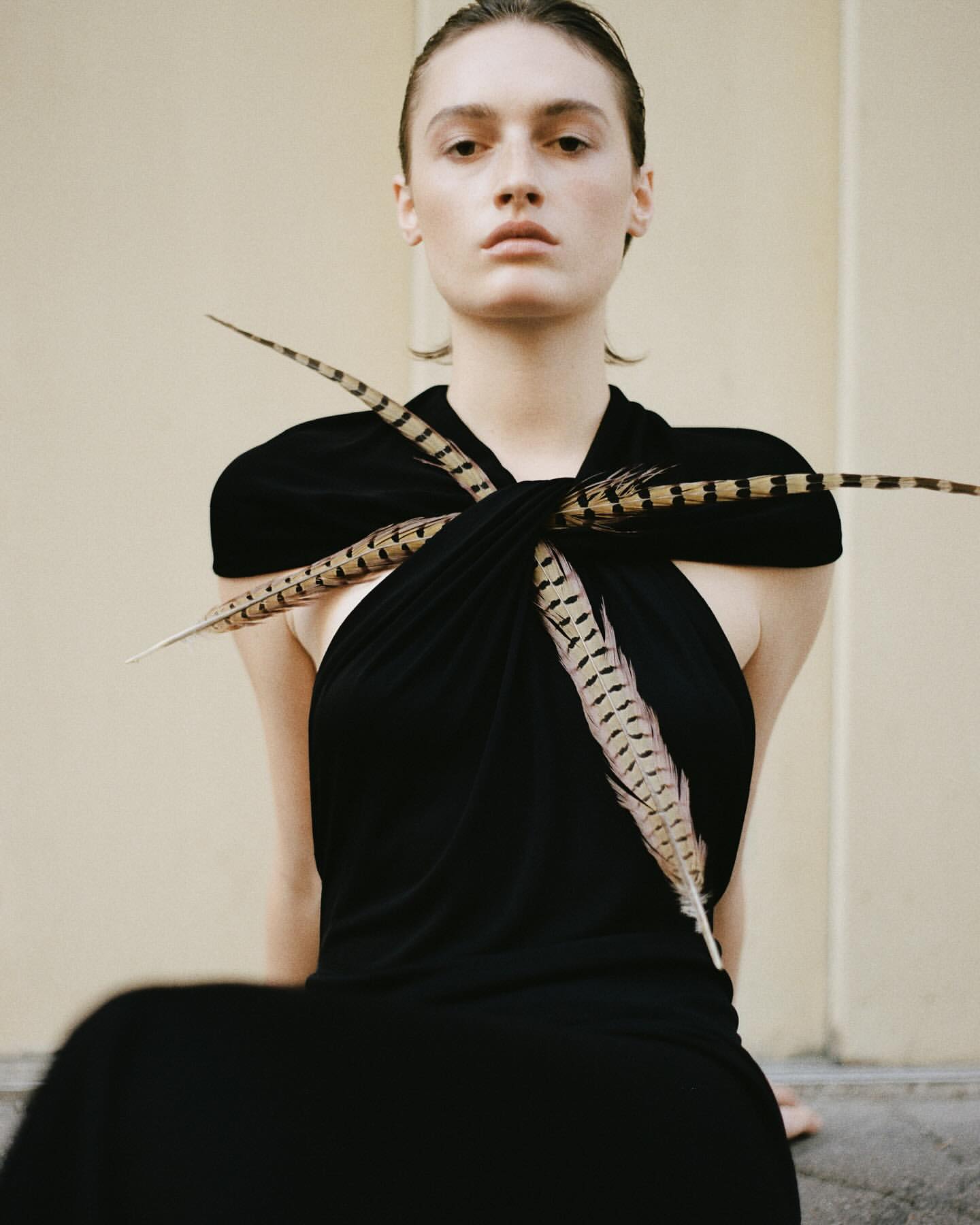 Young woman with fair skin poses in black dress accented with feathers against a light-beige neutral backdrop in a studio shot.