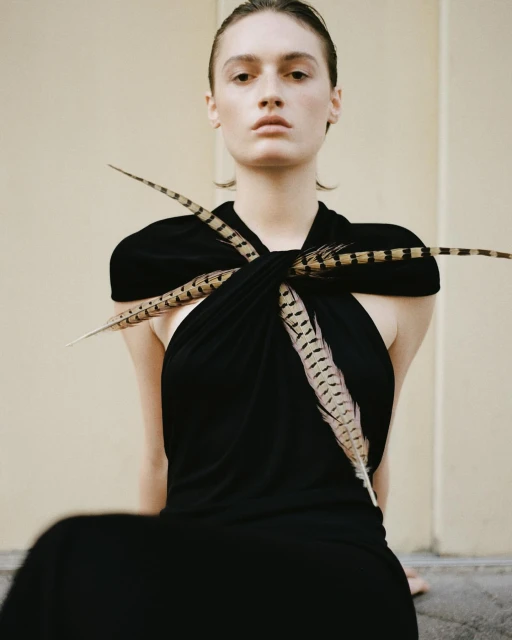 Young woman with fair skin poses in black dress accented with feathers against a light-beige neutral backdrop in a studio shot.