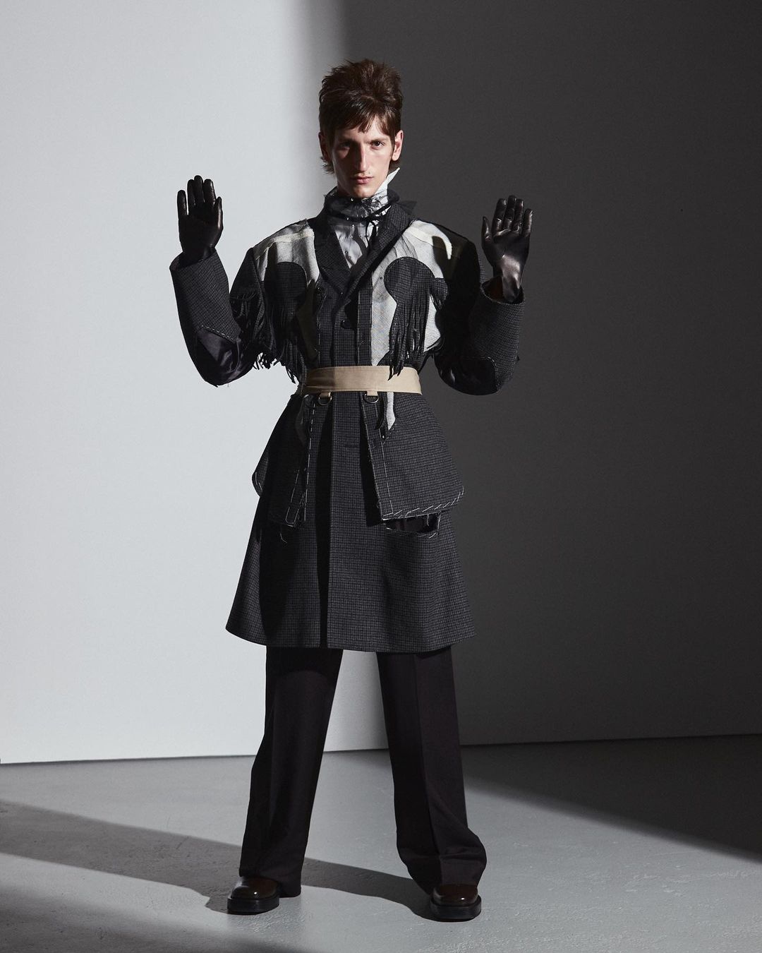 Model wearing a patterned coat with fringe, black trousers, and gloves against a grey studio background.