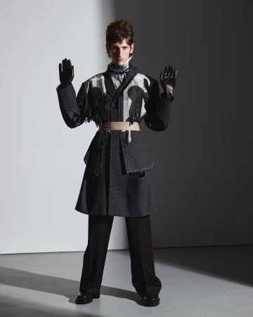 Model wearing a patterned coat with fringe, black trousers, and gloves against a grey studio background.