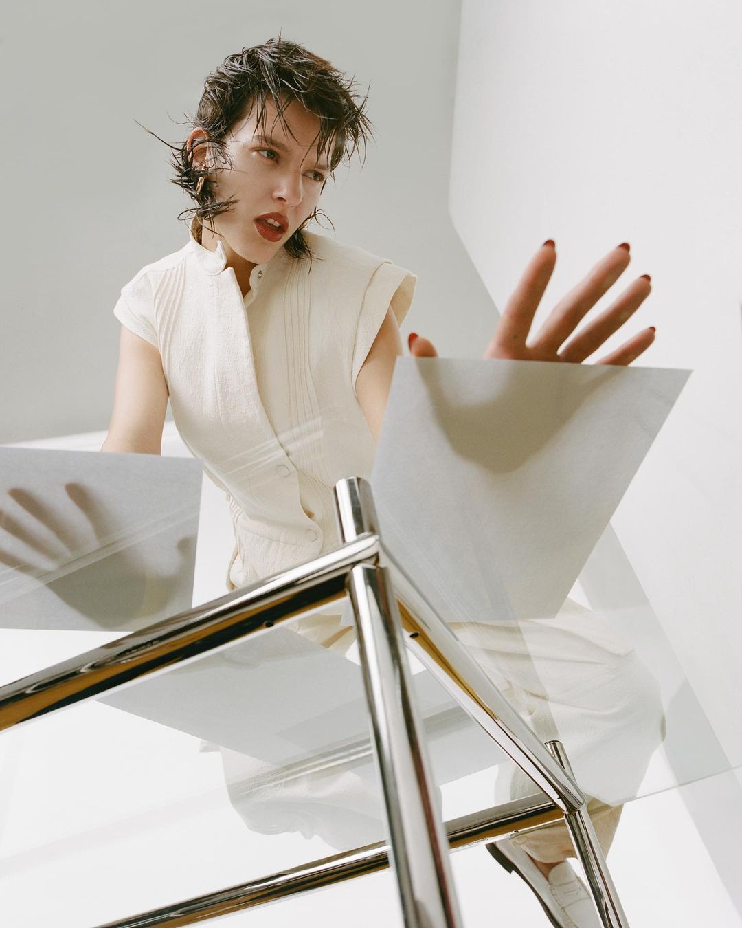 Young woman in white top reaches towards a glass table in a bright, minimalist studio setting, shot from a low angle.