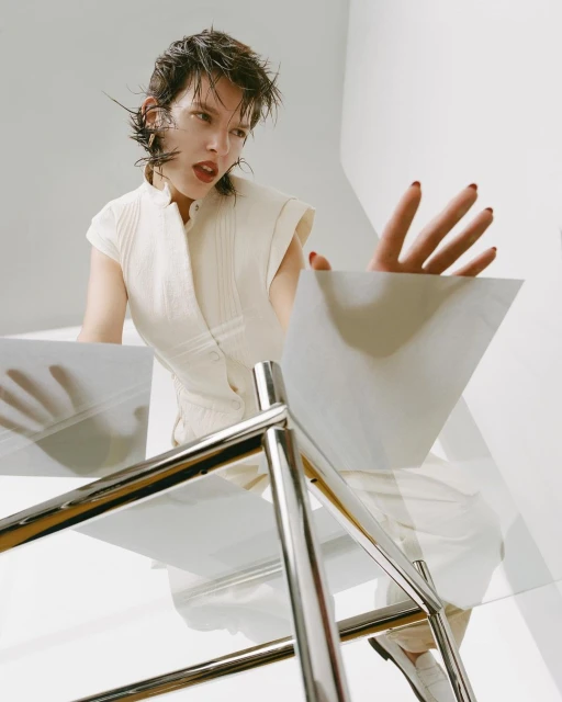 Young woman in white top reaches towards a glass table in a bright, minimalist studio setting, shot from a low angle.