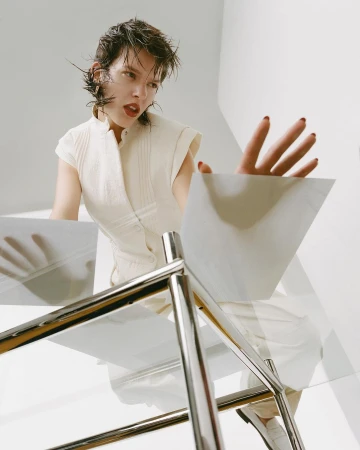 Young woman in white top reaches towards a glass table in a bright, minimalist studio setting, shot from a low angle.