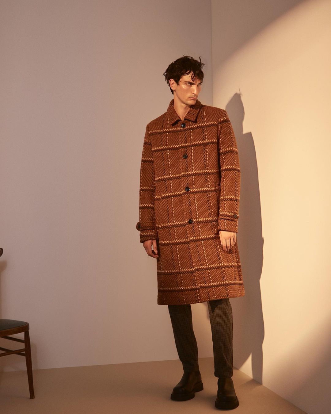 Man wearing a brown patterned overcoat standing in a studio with harsh side lighting and a visible wall shadow.