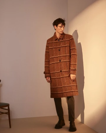 Man wearing a brown patterned overcoat standing in a studio with harsh side lighting and a visible wall shadow.