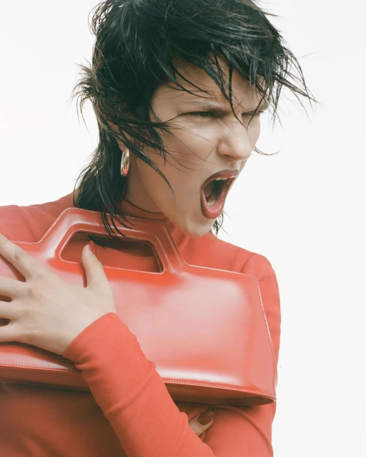 An adult woman with an open mouth and a red top is holding a shiny red handbag in a studio setting with a light background.