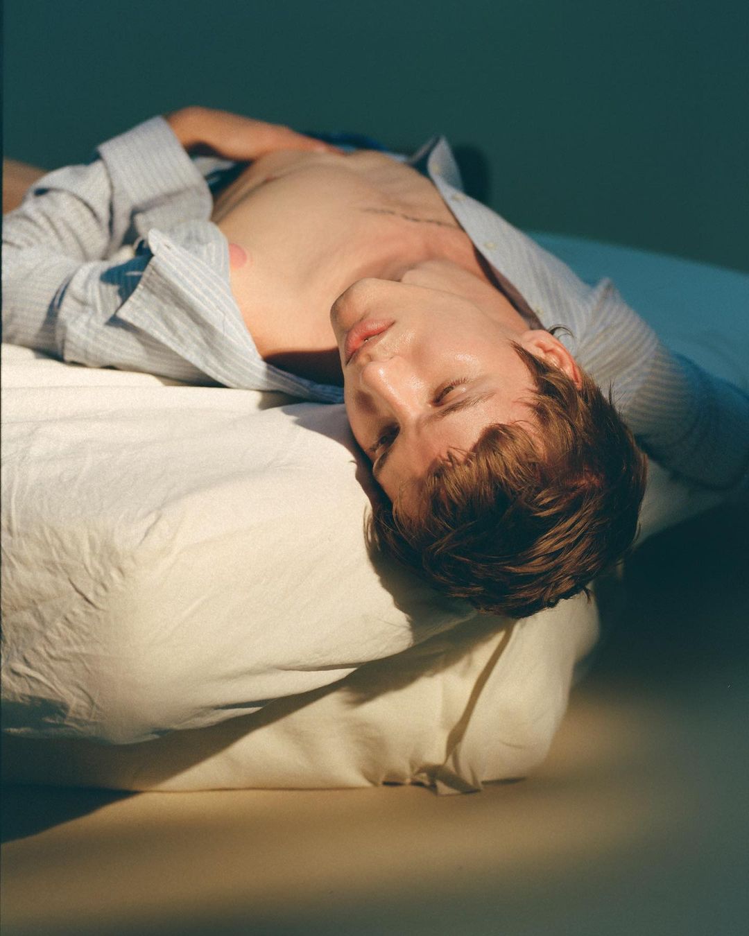 Indoor studio portrait of a young adult male lying on a white pillow with a light blue striped shirt and a calm facial expression.