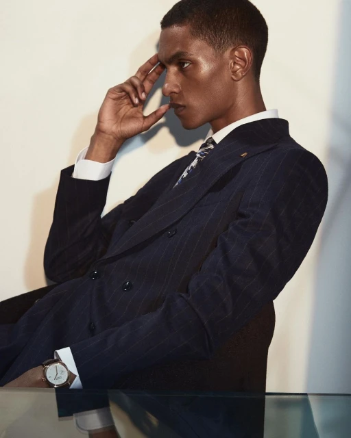 Fashionable adult man in a pinstripe suit poses indoors with a wristwatch and contemplative expression, set against a neutral backdrop.