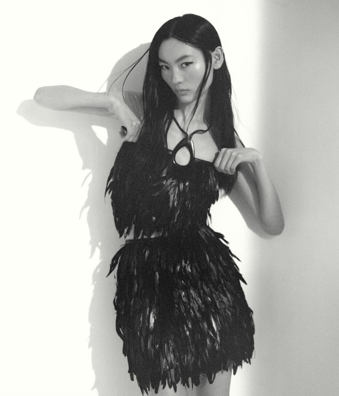 An Asian woman with long black hair poses in a black feather dress and pendant against a white wall with her shadow visible.