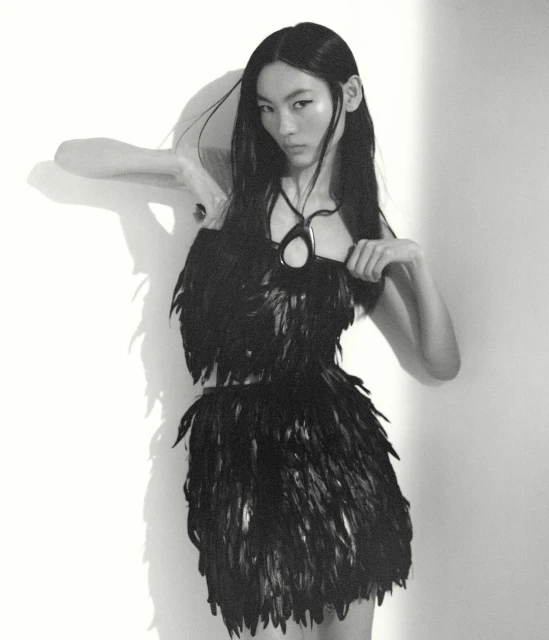 An Asian woman with long black hair poses in a black feather dress and pendant against a white wall with her shadow visible.