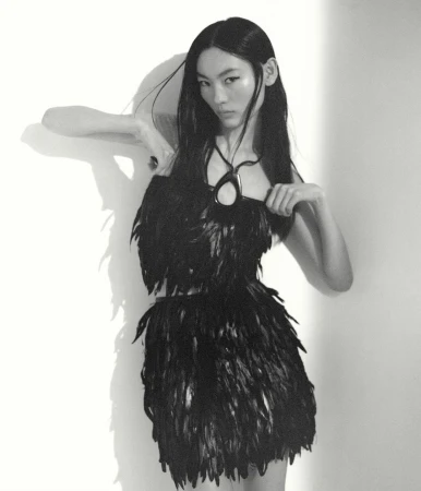 An Asian woman with long black hair poses in a black feather dress and pendant against a white wall with her shadow visible.