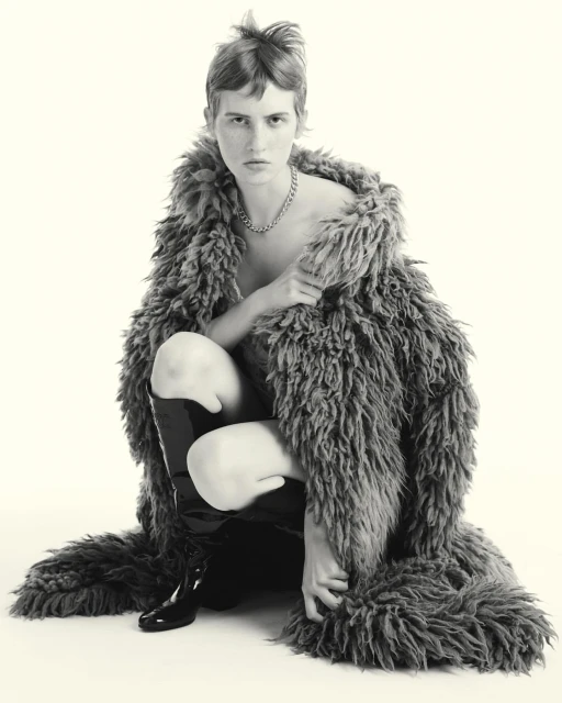 Monochrome fashion shot of a person crouching in a faux fur coat with shiny black boots against a white backdrop, contemplative pose.