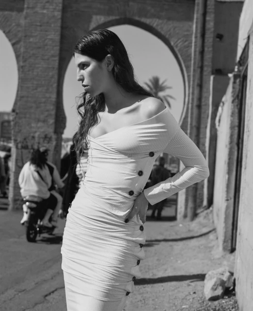 Black and white medium shot of a woman in a fitted dress standing in an arched outdoor structure with a motor scooter in the background.